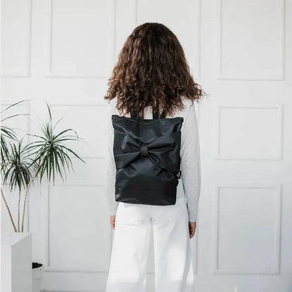 A woman with long curly hair wearing a black backpack featuring a large bow. Versatile and waterproof, perfect for work or everyday use, with adjustable straps and multiple pockets for organization.