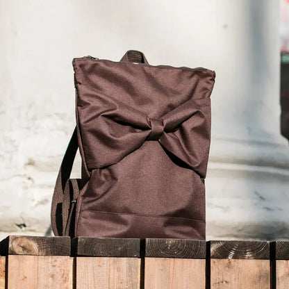 Brown waterproof MUNI backpack with a bow detail, ideal for daily use. Features zipper closure, laptop pocket, and adjustable straps for comfort. Dimensions: 38cm height, 34cm width.