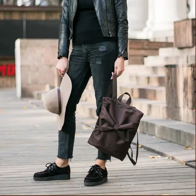 A person holding a brown bow backpack, featuring waterproof brown polyester with a large bow detail. Roomy with laptop pocket and adjustable straps for comfort.