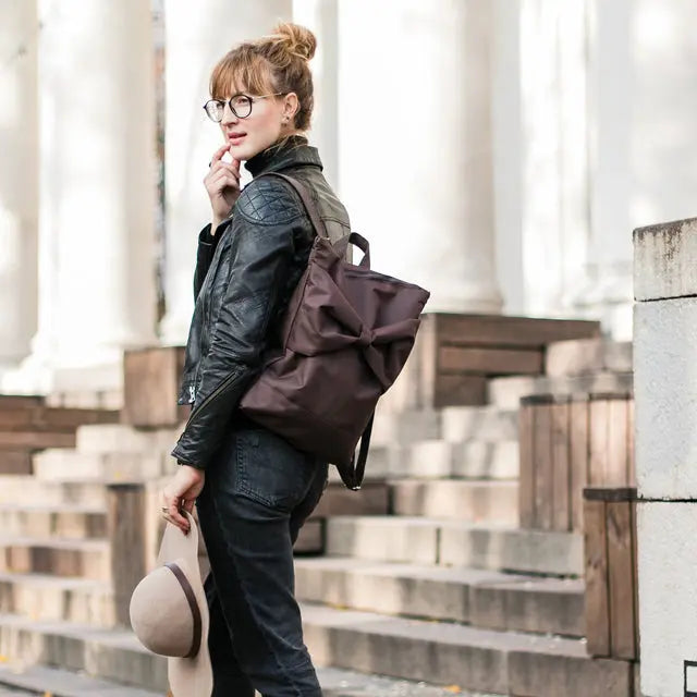 A woman in a leather jacket wearing a brown bow waterproof MUNI backpack, featuring adjustable straps, zipper closure, and laptop pocket.