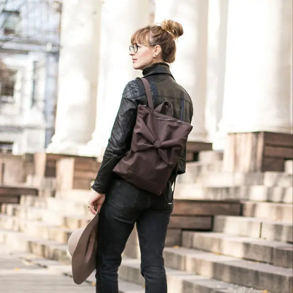 A woman in a leather jacket stands on stairs with a brown Bow backpack. Waterproof polyester material, adjustable straps, zipper closure, laptop pocket. Height: 38 cm, Width: 34 cm.